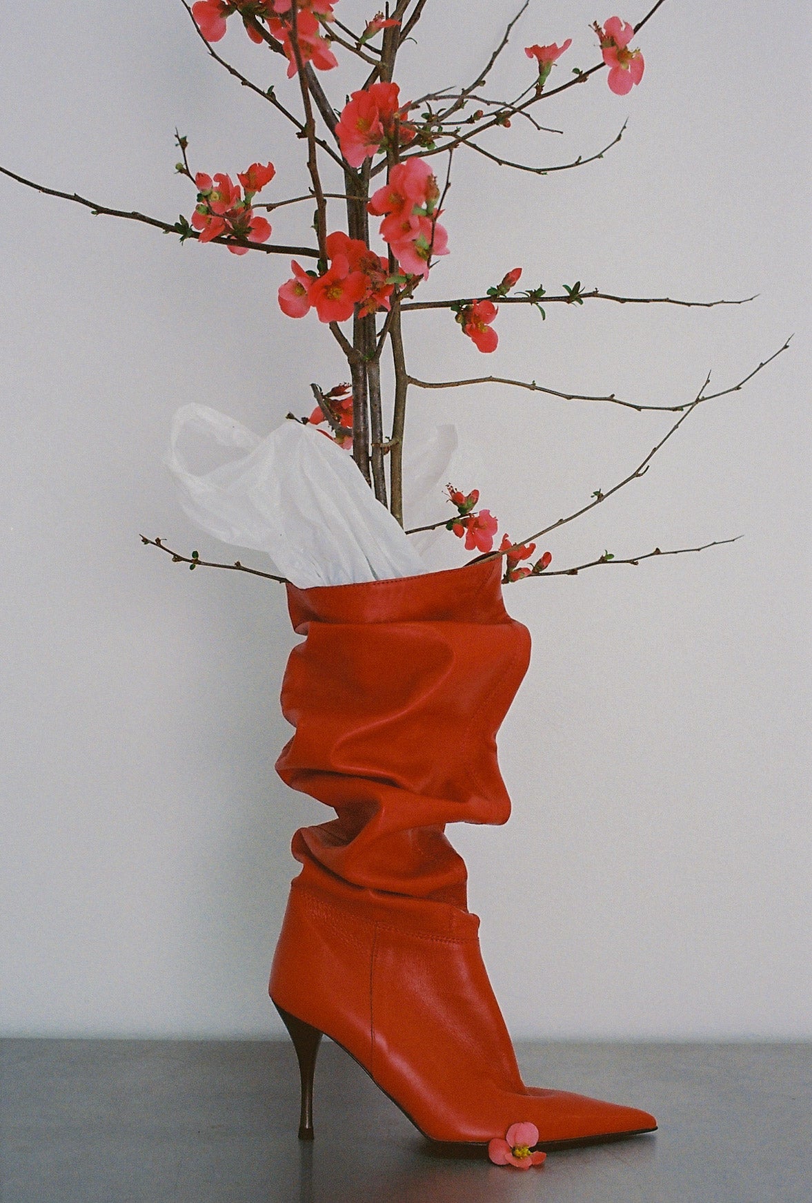 Red high-heeled boots with a white bag and pink flowers on a gray surface.